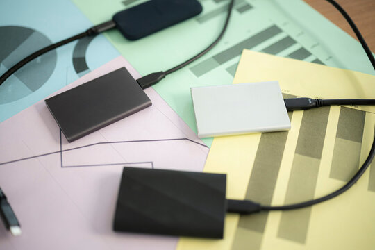 Portable external hard drives connected with black cables rest on colorful business charts and graphs. This modern office workspace setup highlights digital data storage.