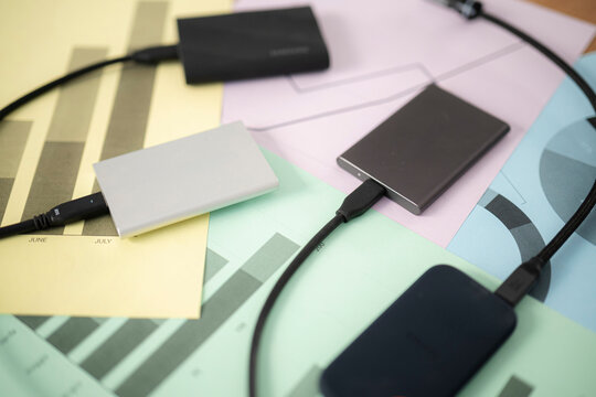 Several portable external solid-state drives are connected with black USB cables and arranged neatly on top of colorful business reports and financial charts.