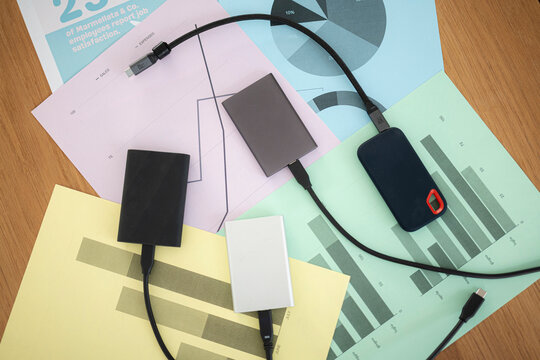 Several external storage drives with connected cables are placed on top of data analysis charts and business reports on a wooden desk during a professional data review.