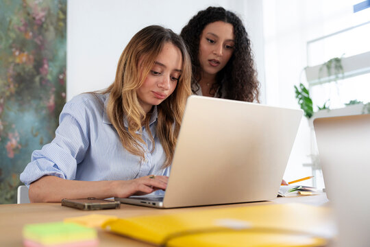 Two young female colleagues collaborate on a project in a bright modern office. One woman types on a laptop while her coworker looks at the screen and gives input.