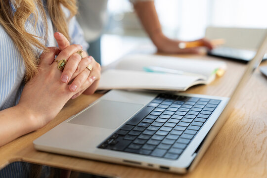 A young woman sits at a wooden office desk with her hands clasped, working on a laptop. Open notebooks and office supplies rest on the table during a productive workday.