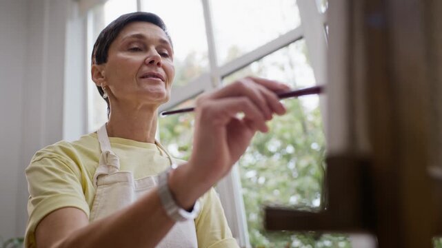 Senior Caucasian woman painting on canvas easel in art class workshop. Footage suitable for promoting creative retirement hobbies, lifelong education, or adult wellbeing programs