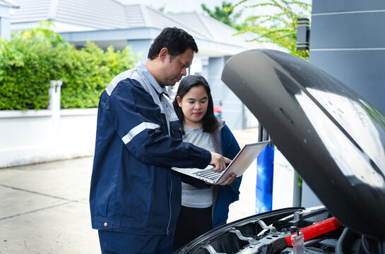 Asian mechanic repairing engine with diagnostic laptop, automotive technician professional diagnosing car breakdown for female customer in driveway of residential garage