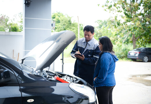 Asian mechanic inspecting car engine and discussing maintenance cost with female vehicle owner at residential driveway for automotive repair service