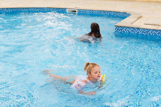 Little blonde girl with floaties swimming in a clear blue outdoor pool while another child swims nearby.