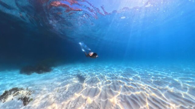 Graceful freediver gliding effortlessly over sandy ocean floor wearing long white fins in crystal clear blue sea. Underwater tracking shot.