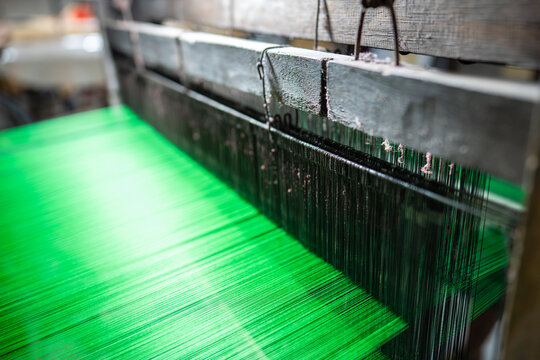 Green threads stretched on traditional wooden loom in textile workshop, traditional fabric manufacturing, colorful yarn for textile production at authentic looming machine, working process at factory.