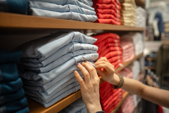 Woman purchasing new basic clothing at garment fashion showroom, woman looking through stack of t-shirts on shelf display, examinees quality of material, fabric and preferable size of clothing item.