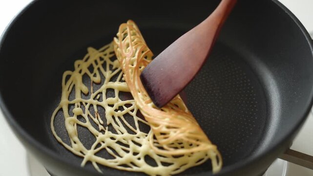 Rolling a crispy lace crepe made from drizzled batter in a nonstick pan with a wooden spatula