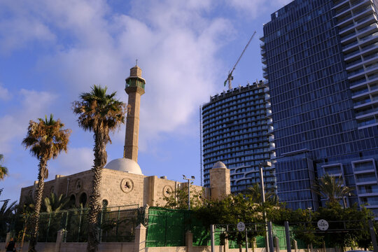 In Tel Aviv, Israel, a traditional mosque stands alongside tall, modern skyscrapers under a cloudy sky.