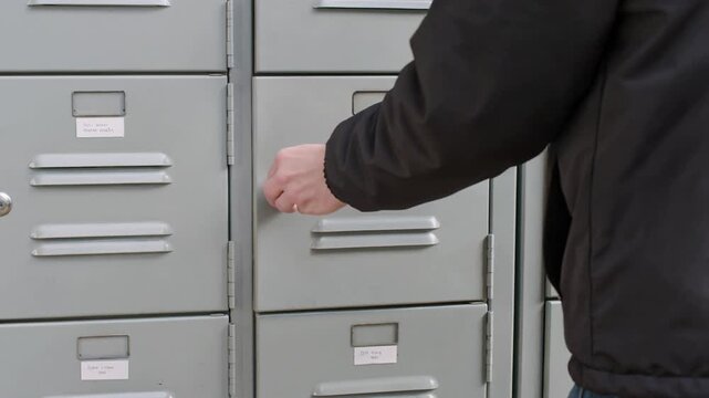 A person in a dark jacket places a cardboard box on a shelf beside gray metal lockers in an indoor storage area