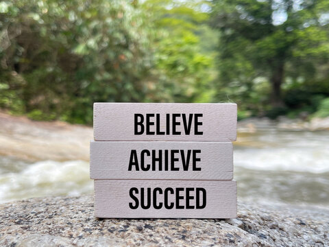 Inspirational motivational message reading Believe Achieve Succeed displayed on stacked wooden blocks beside a flowing river, symbolizing ambition, determination, and personal growth.