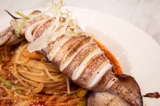 Grilled whole squid spaghetti pasta with tomato sauce on white plate.
