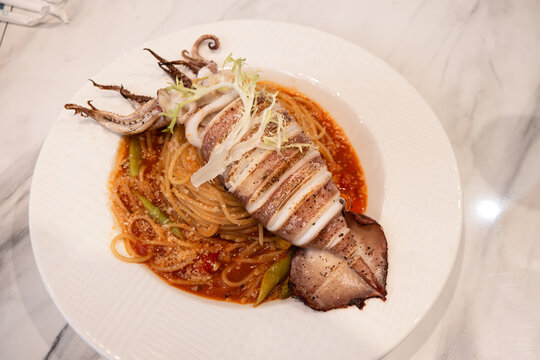 Grilled whole squid spaghetti pasta with tomato sauce on white plate.
