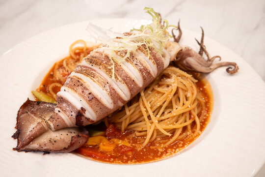 Grilled whole squid spaghetti pasta with tomato sauce on white plate.