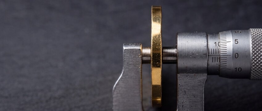 Analog outside micrometer measuring the precise thickness of a gold coin on dark textured slate background with copy space