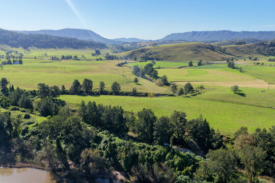 Aerial view across green farmland and rolling wooded hills in Lambs Valley, with a tree lined waterway and rural properties in the Hunter Valley, New South Wales, Australia.