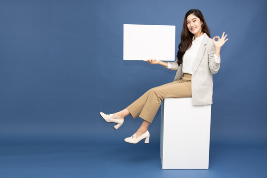 Smiling Asian businesswoman holding a blank white sign and showing OK hand sign on blue background, presenting commercial copy space for marketing promotion, banner, and business advertisement