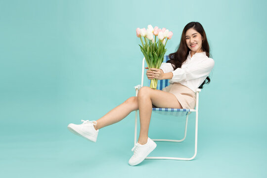 Beautiful Asian woman holding tulip flowers bouquet sitting on chair against mint green background, celebrating Mother's Day, Valentine's Day, spring season, love, romance, and happy lifestyle