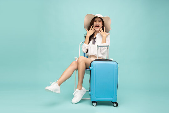 Excited Asian female traveler wearing hat sitting on chair with blue suitcase isolated on mint green background, laughing with joy for summer vacation holiday, journey, and tourism concept