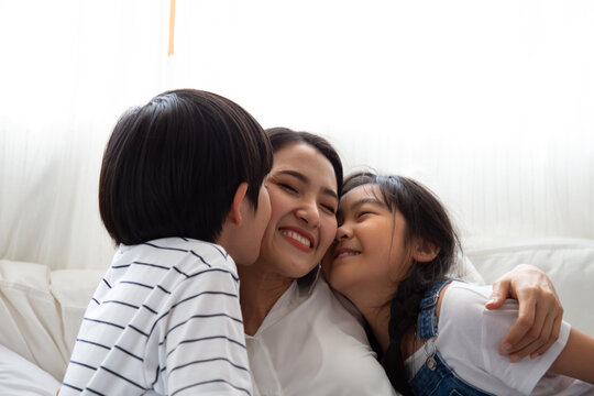 Loving Asian son and daughter kissing and hugging their happy mother on cheek at home, celebrating Mother's Day, family togetherness, childhood affection, care, and sweet home relationship concept