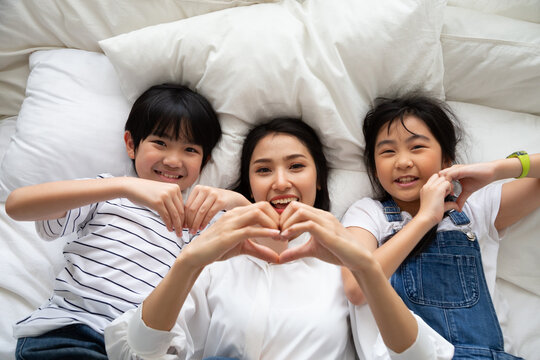 Top view of Asian mother, son, and daughter lying on bed together making heart hand gesture to celebrate Mother's Day, family love, childhood happiness, togetherness, and warm home relationship