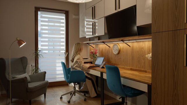 Modern home office room features custom built-in cabinetry, television, and a double wooden desk. Woman works on a laptop in comfortable apartment living space.