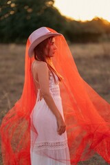 Art portrait, boho wedding, conceptual fashion, setting sun, red veil. Young woman in a white lace dress, hat, and long red veil posing outdoors at sunset, side view profile portrait. © Kateryna