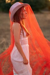Art portrait, boho wedding, conceptual fashion, setting sun, red veil. Young woman in a white lace dress, hat, and long red veil posing outdoors at sunset, side view profile portrait. © Kateryna
