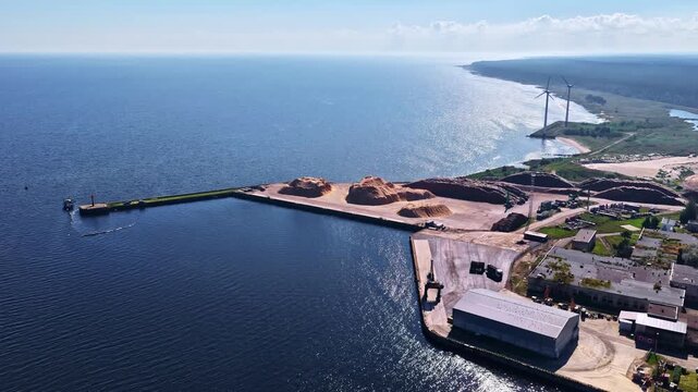 Aerial view of Engure port featuring industrial wood chip piles, storage buildings, and a breakwater extending into the Baltic Sea in Engure, Tukums Municipality, Latvia.