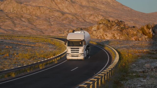 A white tanker truck drives toward the camera along a winding two-lane asphalt road, framed in a compressed telephoto view against sunlit rocky desert hills.