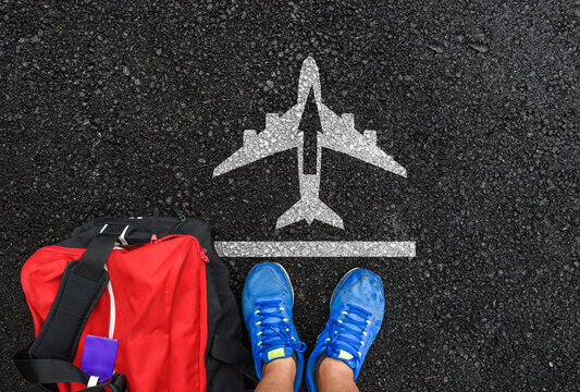 Top down view of person feet with suitcase standing on asphalt road with painted airplane silhouette and arrow, ready for travel and new adventure concept.