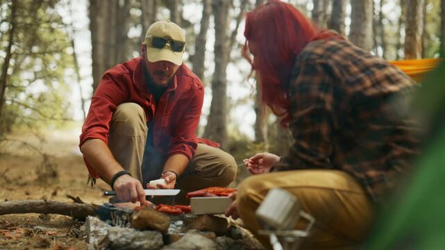 Campers cook food over a campfire in a pine forest, cutting ingredients and tending sausages beside a tent.