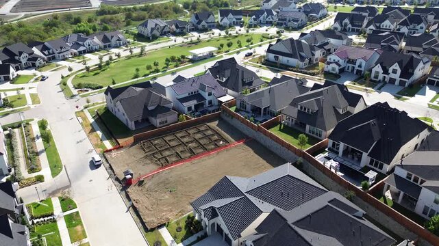 Symmetrical home foundations emerge on cleared parcels bordering occupied family residences along Frisco. Wooden fences outline backyard perimeters along newly developed suburban roadway, Texas