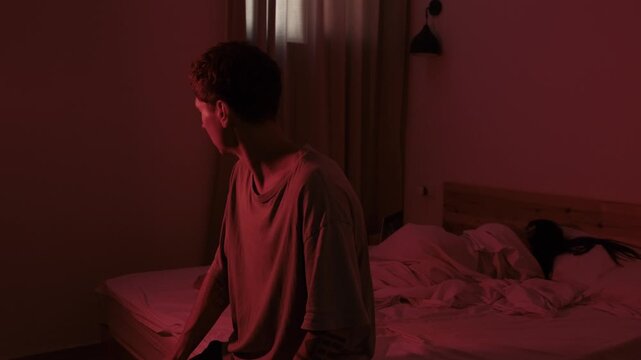Depressed looking man sitting beside his partner while she focuses on a laptop, portraying emotional neglect, lack of intimacy and relationship dissatisfaction in a red lit bedroom