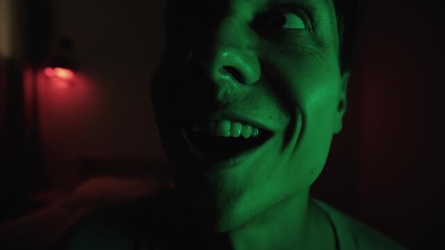 Disturbed man talking to himself in an extreme close up, turning and muttering with an unsettling expression under red and green low key lighting, portraying mental breakdown