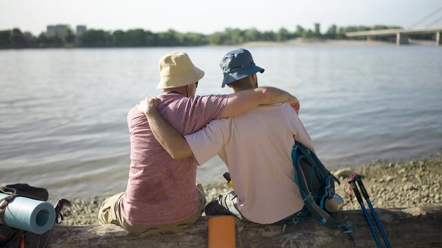 Two male partners lean together beside a lake, enjoying each other's company and the tranquil outdoor scenery.