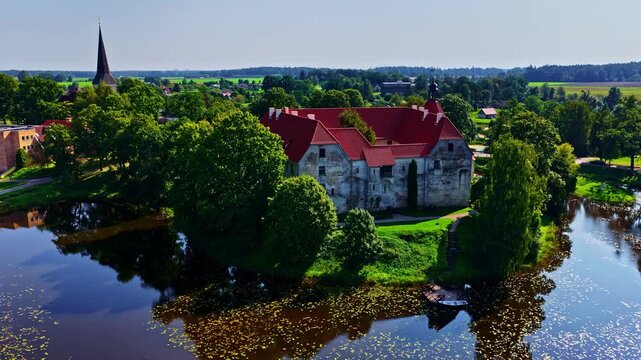 Aerial view of Jaunpils Castle with its distinctive red roof and stone walls, surrounded by a moat and lush greenery in Jaunpils, Tukums Municipality, Latvia.