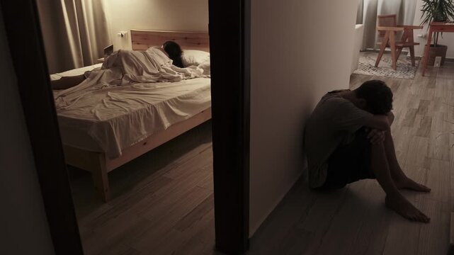 Emotionally withdrawn man sitting on the floor with head down while his partner lies turned away in bed, portraying relationship crisis, silent treatment and emotional pain at home