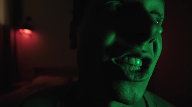 Unhinged man staring and turning in an extreme close up with distorted facial expressions, muttering to himself under dramatic red and green lighting, portraying madness and mental distress