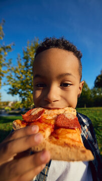 Curly haired boy biting pepperoni pizza in sunny park closeup with delight. Childhood enjoyment, casual dining concept.