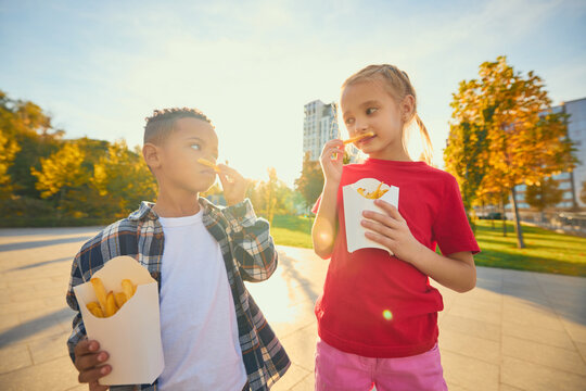 Boy and girl eating fries from takeaway cartons on park path with cheerful interest. Childhood friendship, casual dining concept.