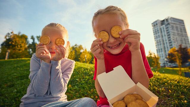 Playful girls holding round crackers over eyes on park grass with laughter. Childhood imagination, social bonding concept.