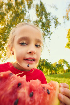 Playful little girl holding bitten watermelon close to camera in sunny park. Childhood joy, fresh nutrition concept.