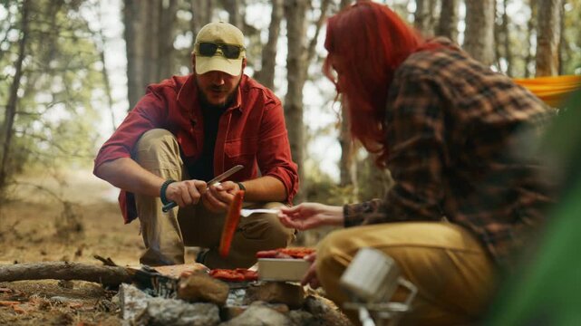 Campers cook food over a campfire in a pine forest, peeling an egg and tending sausages beside a tent.
