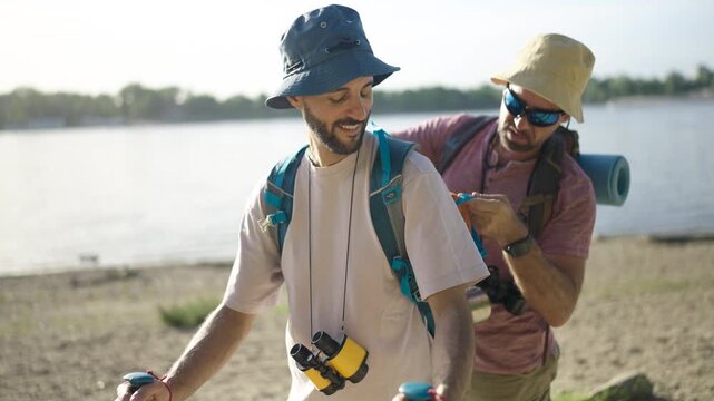 A male couple walks beside the water with hiking equipment, exploring the shoreline during a relaxing outdoor excursion.