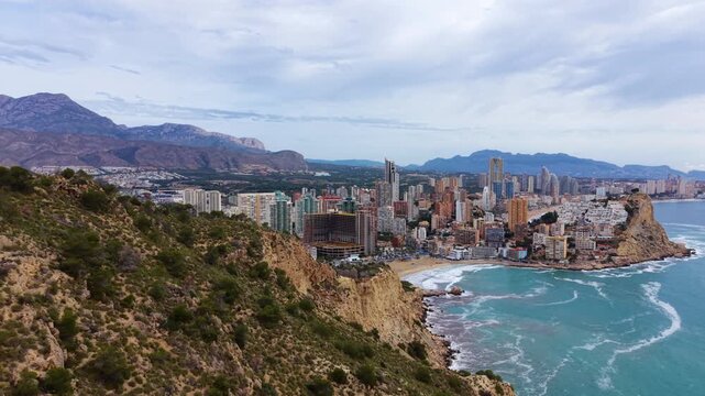 Elevated view of Benidorm beachfront with tall hotel towers, sandy Levante beach, rocky headland and turquoise Mediterranean waves under cloudy sky