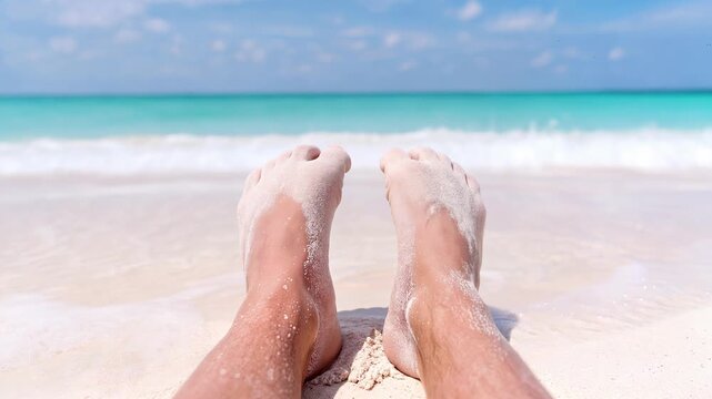 Relaxation and carefree escape found on tropical beach as male sandy feet rest before turquoise ocean waves. Personal perspective view.