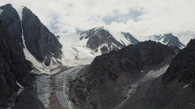 Aerial photograph captures the Aktru Valley glacier, fractured ice and steep stone ridges beneath a shifting cloud layer, revealing the remote alpine landscape of Altai Republic, Russia