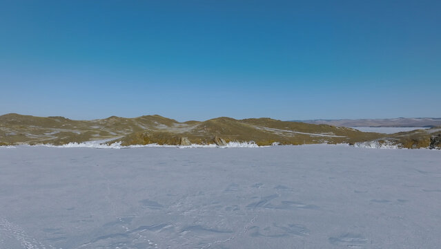 Snow-covered ice spreads across Lake Baikal near the Maloye More strait, bordered by distant hills beneath a clear sky in Irkutsk Oblast, Russia, a broad winter view of remote Siberia